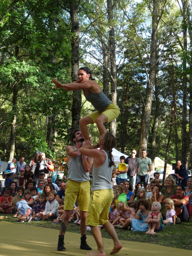 Deux hommes portant une femme accroupie, bras devant elle, devant un public dans un camping